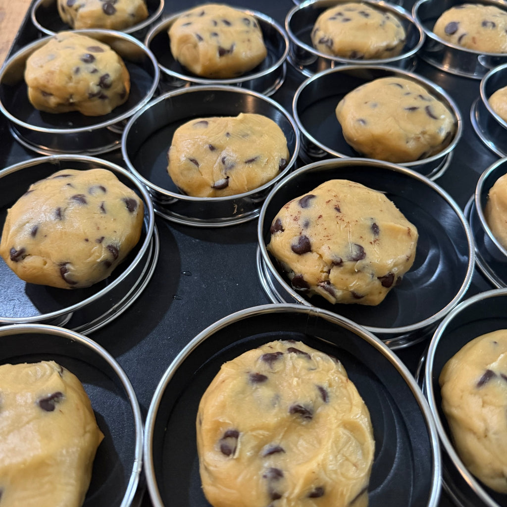 SEBA Artisan Sourdough Chocolate Chip Cookie - Artisan Real Sourdough Bread Chef SebaArtisan Real Sourdough Bread Chef SebaArtisan Real Sourdough Bread Chef SebaSEBA-CCC-120Unbaked chocolate chip cookie dough balls pressed into round metal cookie cutters on a black baking tray, with more trays in the background.
