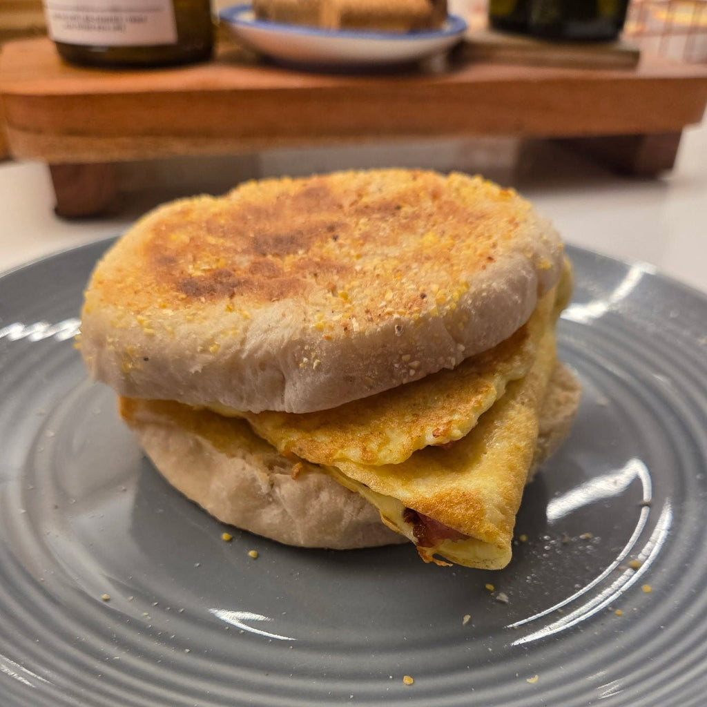 SEBAlicious™ Artisan Sourdough English Muffins Tuesday special - Artisan Real Sourdough Bread Chef SebaArtisan Real Sourdough Bread Chef SebaArtisan Real Sourdough Bread Chef SebaSEBA-EM-4-SD4 packA toasted English muffin sandwich with a folded egg and a small piece of bacon, served on a gray plate in a kitchen setting.