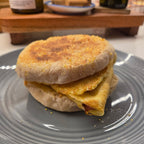 SEBAlicious™ Artisan Sourdough English Muffins Tuesday special - Artisan Real Sourdough Bread Chef SebaArtisan Real Sourdough Bread Chef SebaArtisan Real Sourdough Bread Chef SebaSEBA-EM-4-SD4 packA toasted English muffin sandwich with a folded egg and a small piece of bacon, served on a gray plate in a kitchen setting.