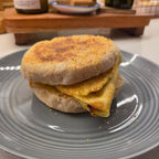 SEBAlicious™ Artisan Sourdough English Muffins Tuesday special - Artisan Real Sourdough Bread Chef SebaArtisan Real Sourdough Bread Chef SebaArtisan Real Sourdough Bread Chef SebaSEBA-EM-4-SD4 packA toasted English muffin sandwich with a folded egg and a small piece of bacon, served on a gray plate in a kitchen setting.