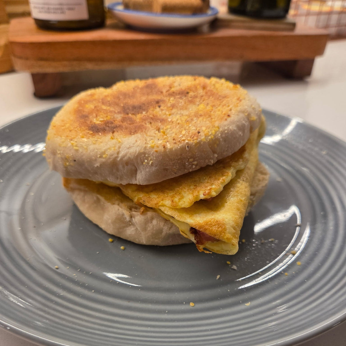 SEBAlicious™ Artisan Sourdough English Muffins Tuesday special - Artisan Real Sourdough Bread Chef SebaArtisan Real Sourdough Bread Chef SebaArtisan Real Sourdough Bread Chef SebaSEBA-EM-4-SD4 packA toasted English muffin sandwich with a folded egg and a small piece of bacon, served on a gray plate in a kitchen setting.