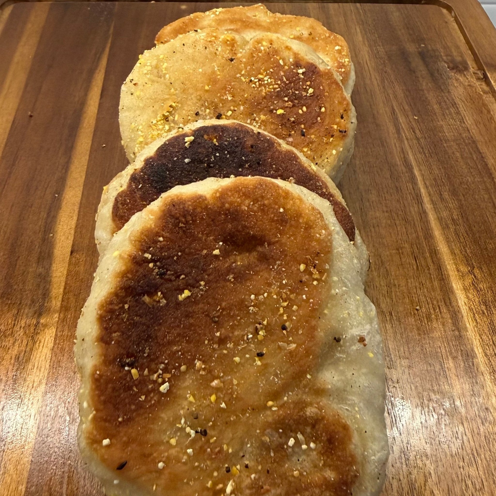 SEBAlicious™ Artisan Sourdough English Muffins Tuesday special - Artisan Real Sourdough Bread Chef SebaArtisan Real Sourdough Bread Chef SebaArtisan Real Sourdough Bread Chef SebaSEBA-EM-4-SD4 packThree golden-brown toasted flatbreads or pancakes sprinkled with herbs, stacked on a wooden cutting board with a white tiled wall in the background.