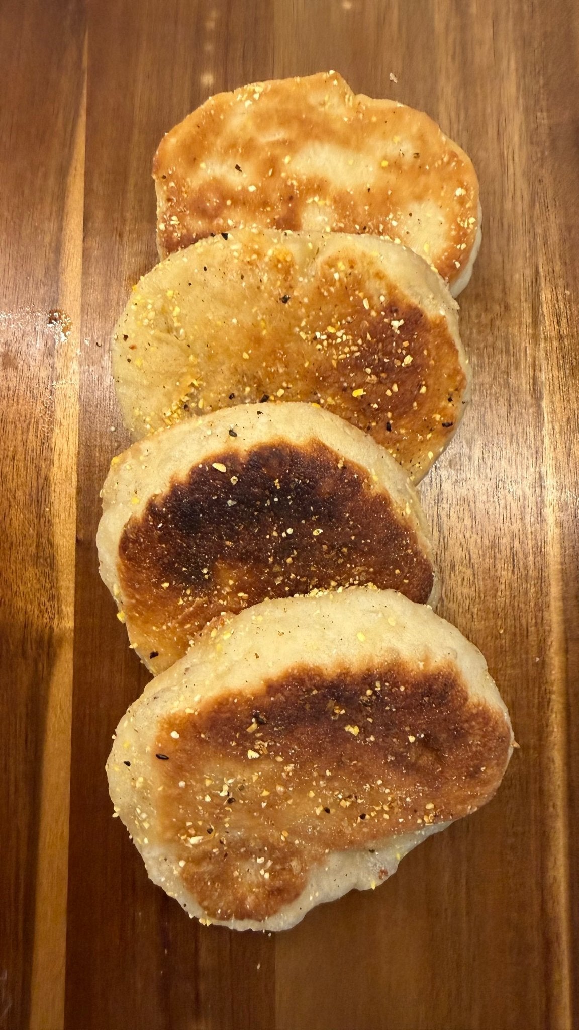 SEBAlicious™ Artisan Sourdough English Muffins Tuesday special - Artisan Real Sourdough Bread Chef SebaArtisan Real Sourdough Bread Chef SebaArtisan Real Sourdough Bread Chef SebaSEBA-EM-4-SD4 packFour toasted English muffins with a sprinkle of seasoning, arranged vertically on a wooden cutting board.