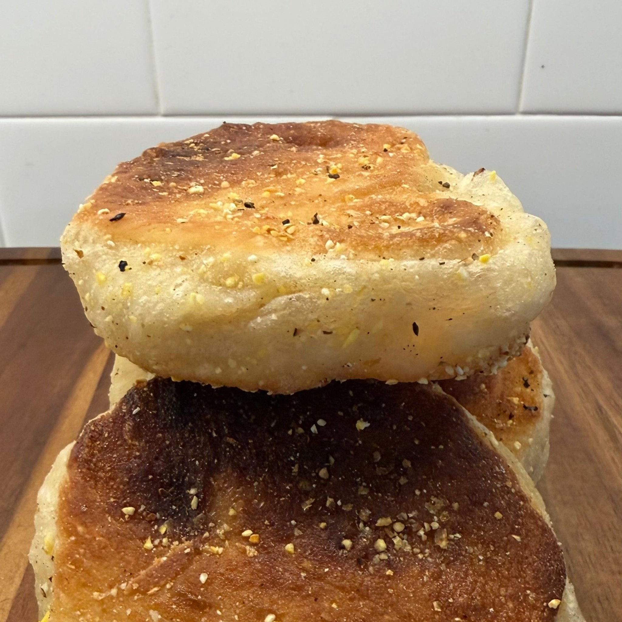 SEBAlicious™ Artisan Sourdough English Muffins Tuesday special - Artisan Real Sourdough Bread Chef SebaArtisan Real Sourdough Bread Chef SebaArtisan Real Sourdough Bread Chef SebaSEBA-EM-4-SD4 packThree toasted English muffins stacked on a wooden cutting board with a white tiled wall in the background.