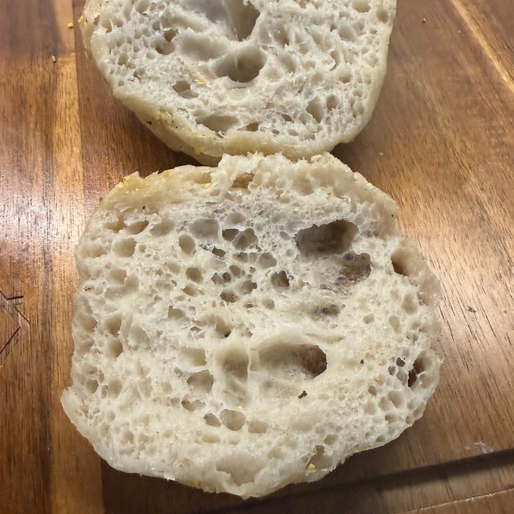 SEBAlicious™ Artisan Sourdough English Muffins Tuesday special - Artisan Real Sourdough Bread Chef SebaArtisan Real Sourdough Bread Chef SebaArtisan Real Sourdough Bread Chef SebaSEBA-EM-4-SD4 packTwo sliced round bread rolls with a golden-brown toasted top, showing a highly porous, airy crumb structure, resting on a wooden cutting board.