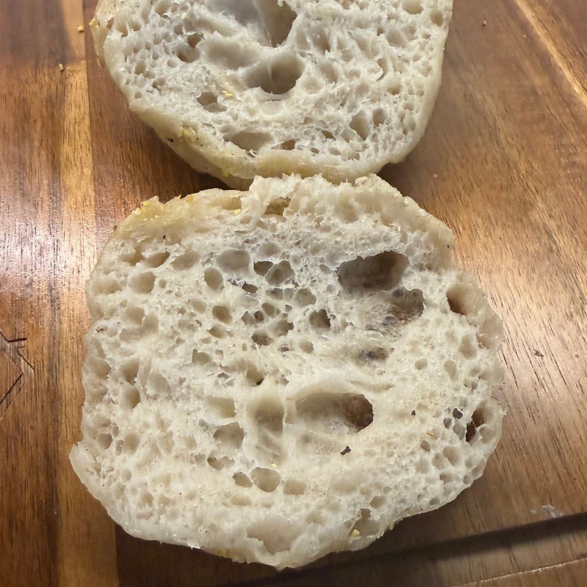 SEBAlicious™ Artisan Sourdough English Muffins Tuesday special - Artisan Real Sourdough Bread Chef SebaArtisan Real Sourdough Bread Chef SebaArtisan Real Sourdough Bread Chef SebaSEBA-EM-4-SD4 packTwo sliced round bread rolls with a golden-brown toasted top, showing a highly porous, airy crumb structure, resting on a wooden cutting board.